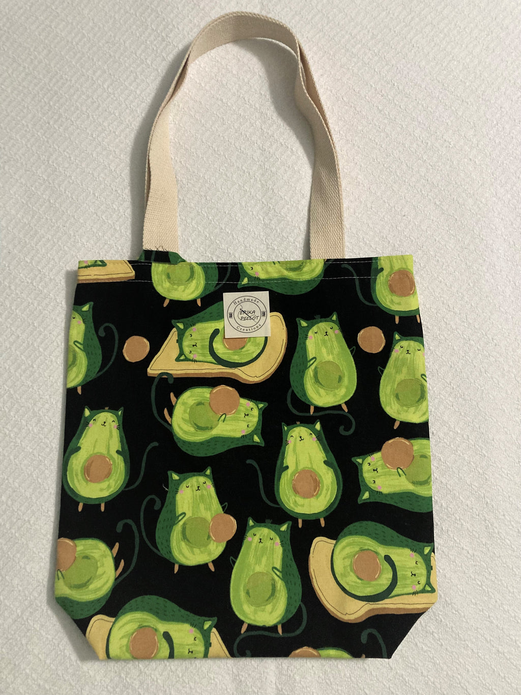 If we mix an avocado and a "gato", we get an AvoGato! This tote has green kitties with a pit for a belly and some are laying down on a piece of bread. This design is in a black background and the tote belting trim handles are of a natural color. All the materials are 100% cotton making it a sustainable item. This cute handmade tote bag is around 15 inches wide and 18 inches long with a 4 inch bottom.