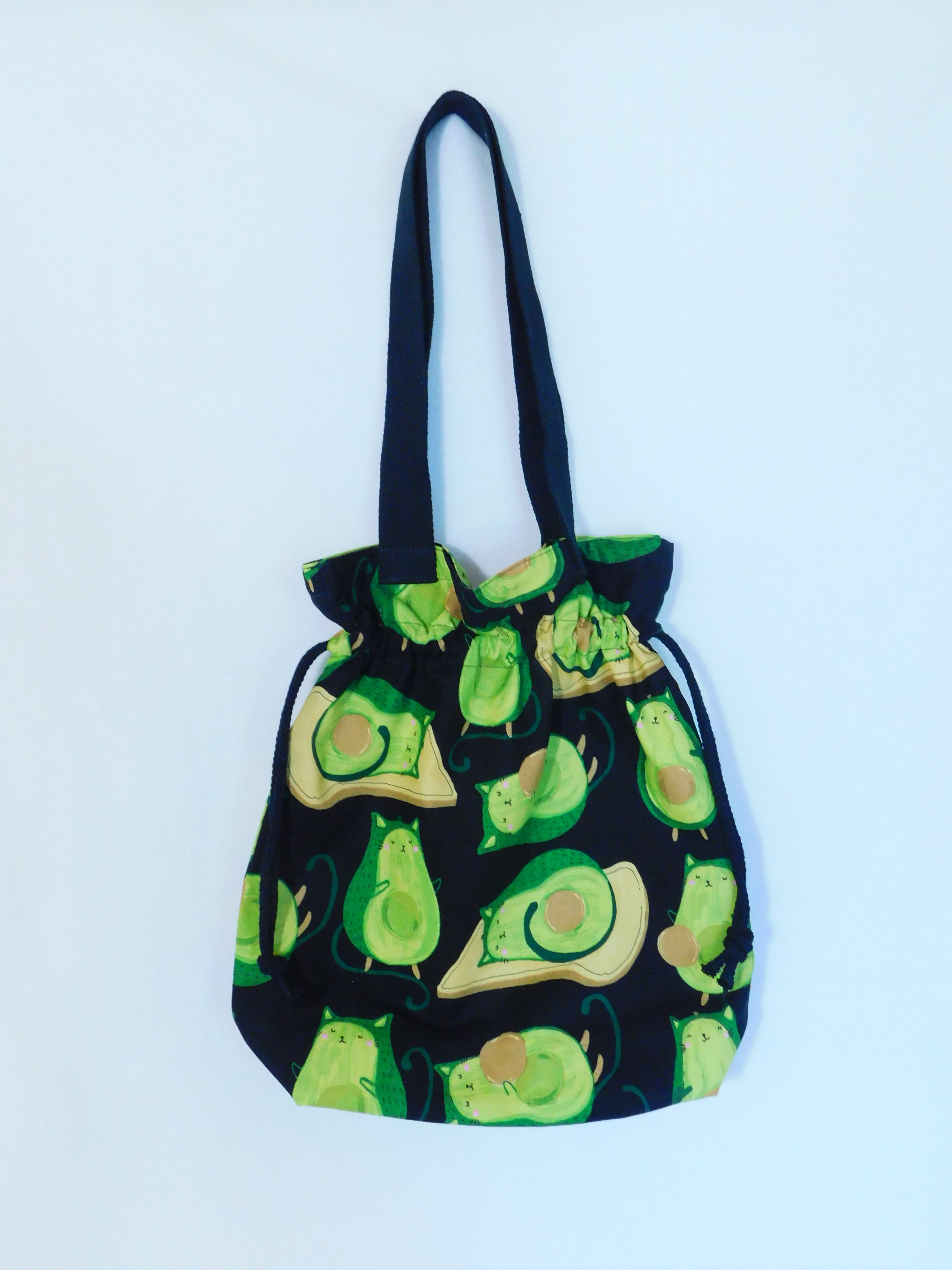 Combine a cat and an avocado and we have a Avo-Gato! This cute tote has cats in the shape of green avocados in a black background and it's just adorable! It has a drawstring to secure your bag and the inside pocket is big enough to fit a cell phone and keys. Fabric, muslin lining, thread and trim are 100% cotton. To care for your tote machine wash gentle cold, tumble dry low, non-chlorine bleach and cool iron. Measurements are around 16in x 14 1/2in x 4in.